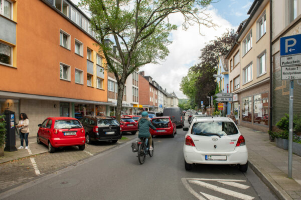 Straße in Werden mit parkenden Autos auf beiden Seiten und einem Radfahrer auf der Fahrbahn, begleitet von Fußgängern und Hinweisschildern zu Parkplätzen.