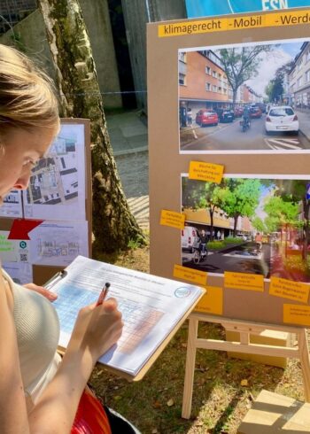 Frau füllt ein Formular aus, während sie vor einer Pinnwand mit Plänen und Visualisierungen für ein klimagerechtes Mobilitätskonzept im Stadtteil Werden steht.