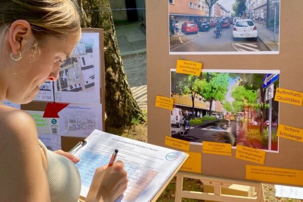 Frau füllt ein Formular aus, während sie vor einer Pinnwand mit Plänen und Visualisierungen für ein klimagerechtes Mobilitätskonzept im Stadtteil Werden steht.